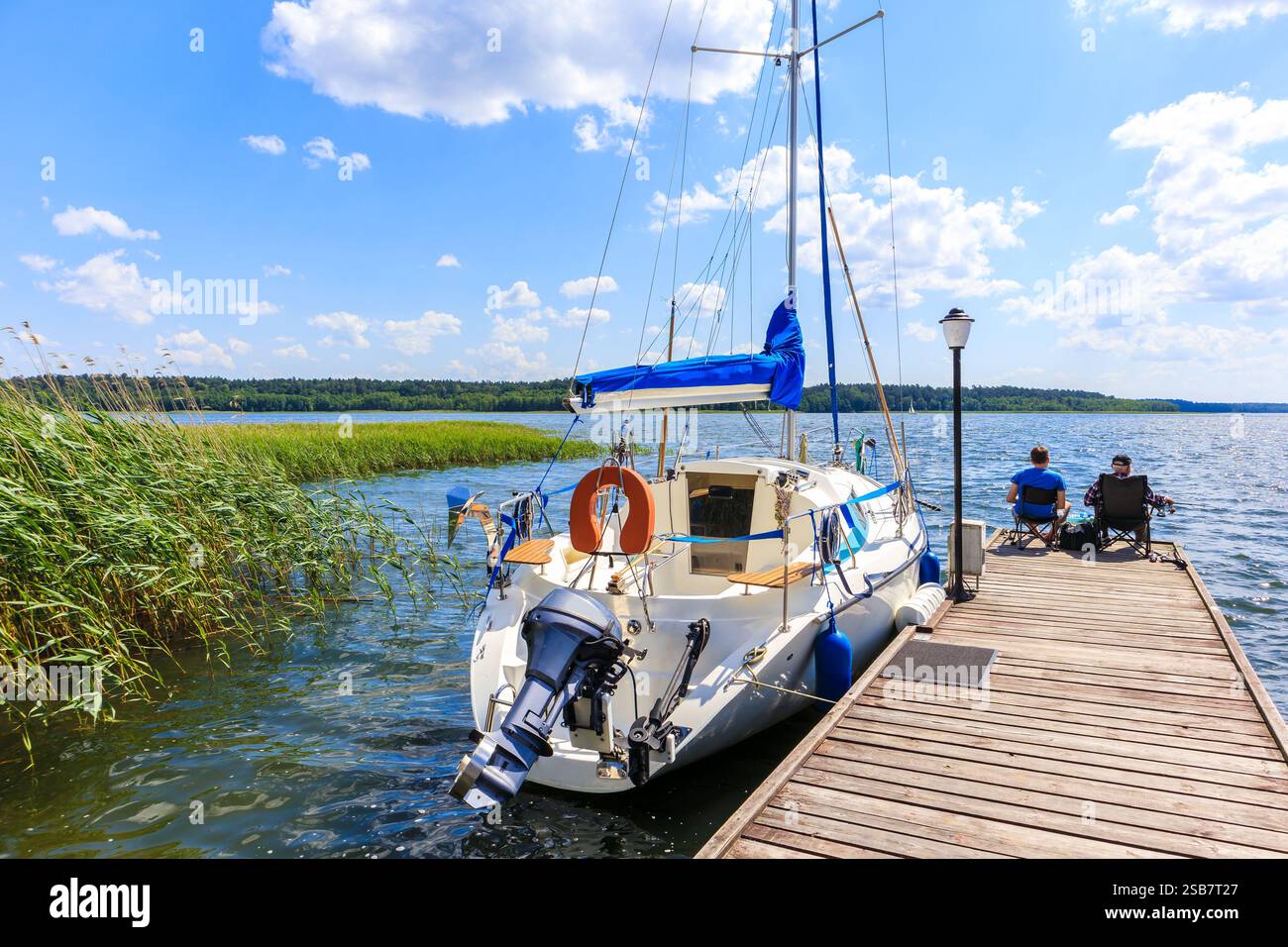 Zwei nicht identifizierte Männer fischen am Pier und Segelboote, die am Seeufer im Hafen von Krzyze an sonnigem Sommertag im Mazury Lake District, Polen, anlegen Stockfoto