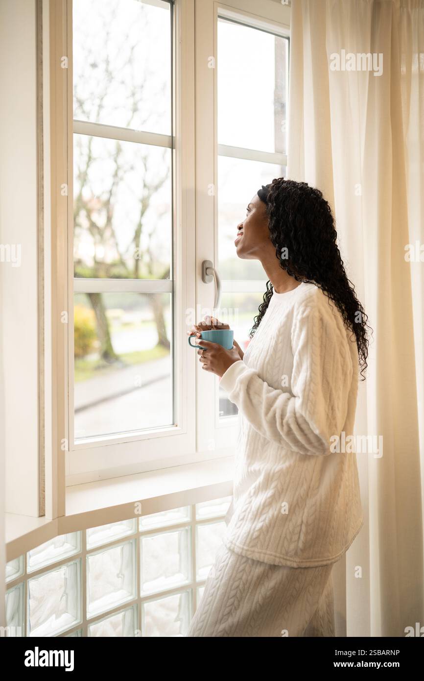 Eine junge Frau in bequemer Loungewear, die einen Becher hält, steht an einem hellen Fenster und blickt nachdenklich auf die Landschaft im Freien, während sie ihren Morgen genießt Stockfoto