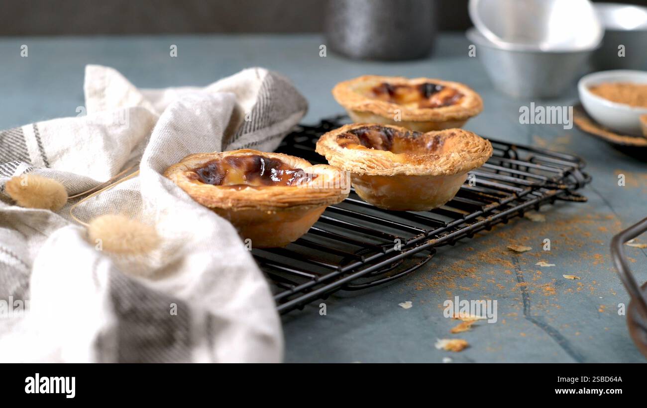 Drei portugiesische Puddingkuchen, die auf einem schwarzen Gestell mit einem gefalteten weißen Zimt und Backformen im Hintergrund abkühlen Stockfoto