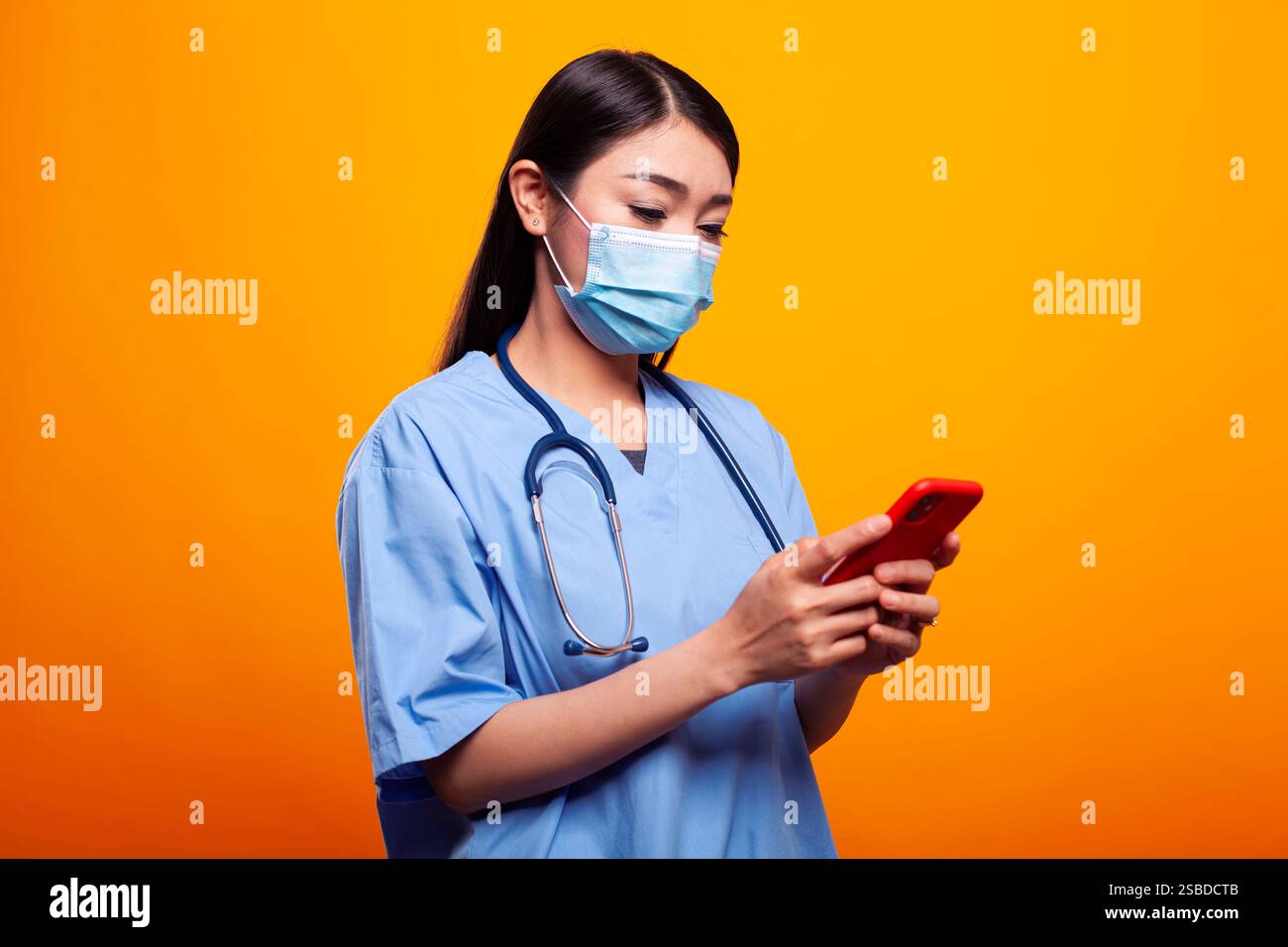Junge Krankenschwester in medizinischer Uniform und Gesichtsmaske, SMS auf ihrem mobilen Gerät, während sie vor isoliertem Hintergrund steht. Asiatische Ärztin mit einem Stethoskop, mit Handy, in ihren sozialen Medien. Stockfoto