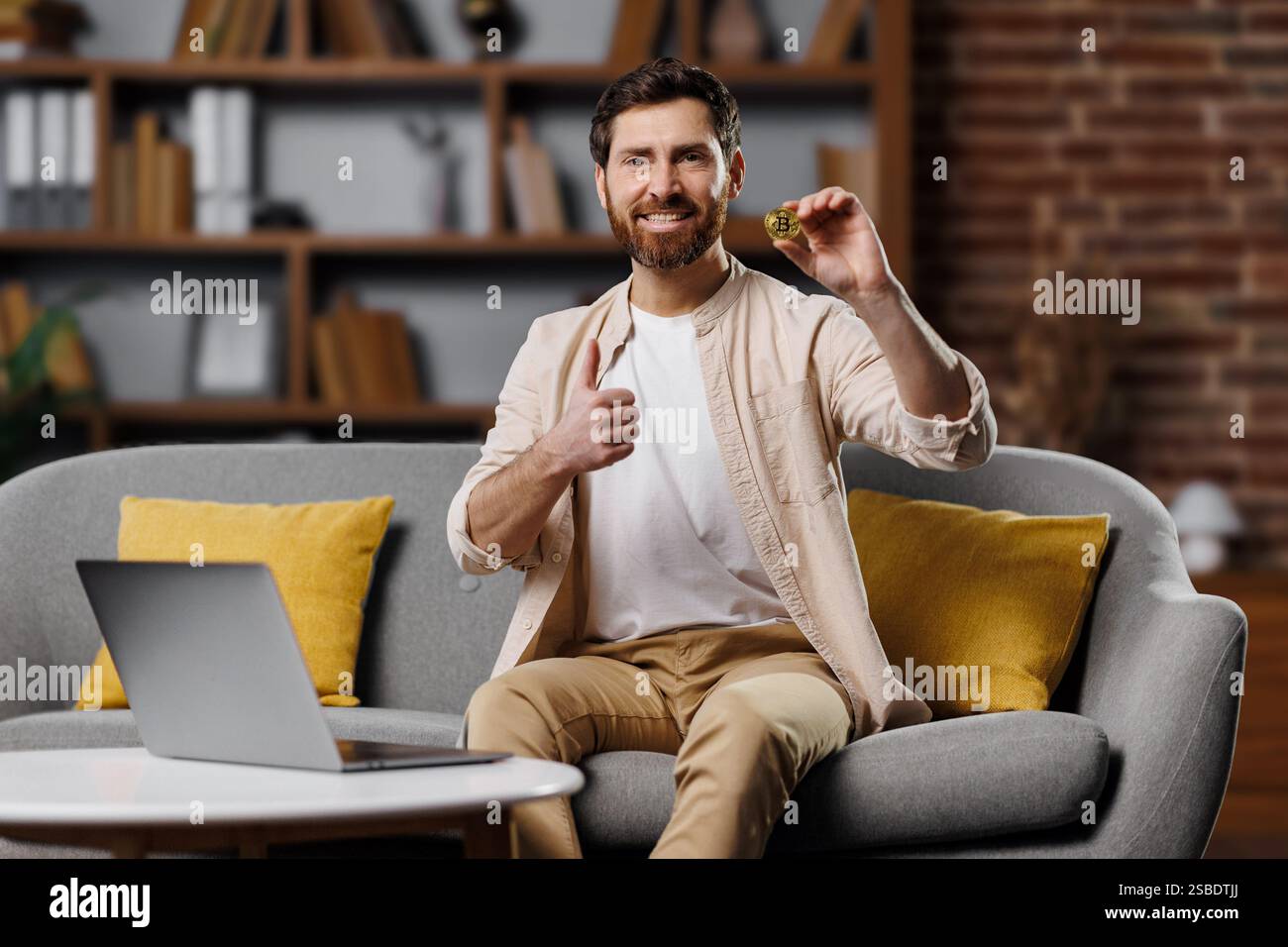 Der Mann lächelt, während er einen Keks hält und in einem gemütlichen Wohnzimmer mit offenem Laptop und heller Einrichtung die Daumen nach oben drückt, was auf einen freudigen Moment hinweist Stockfoto