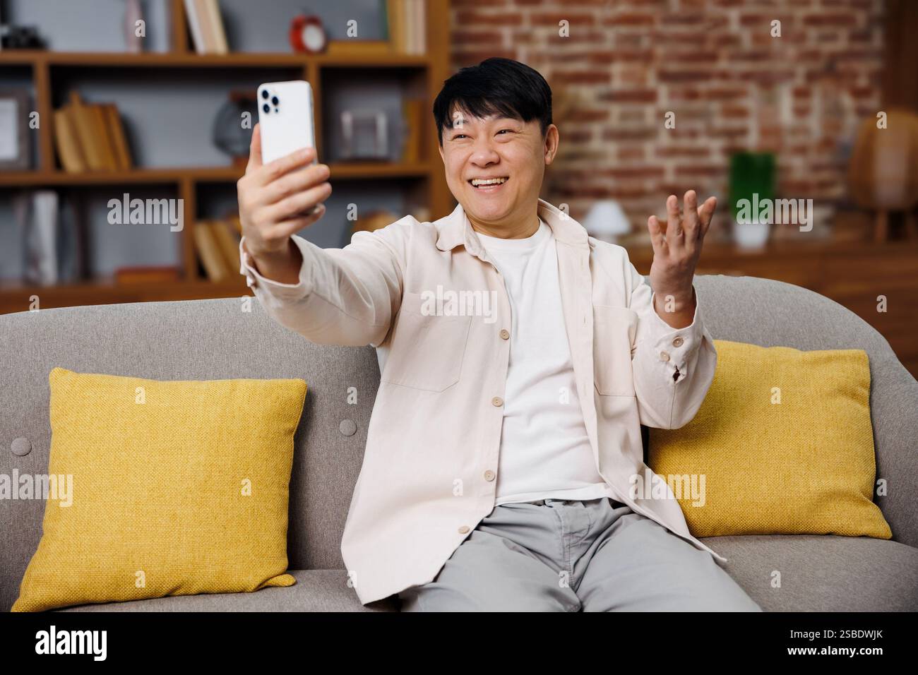 Videogespräche, Telefongespräche, lächelnder asiatischer Mann, genießt Videoanrufe, während er auf einer bequemen Couch in einem gemütlichen Wohnzimmer sitzt Stockfoto