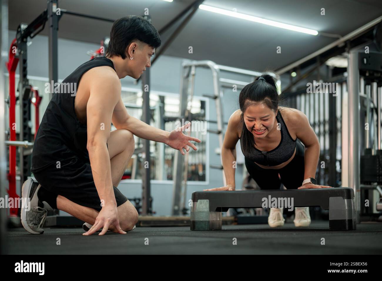 Eine konzentrierte und entschlossene asiatische Frau wird von einem professionellen Fitnesstrainer sorgfältig trainiert, während sie Liegestütze mit einem Schritt und beim Training durchführt Stockfoto