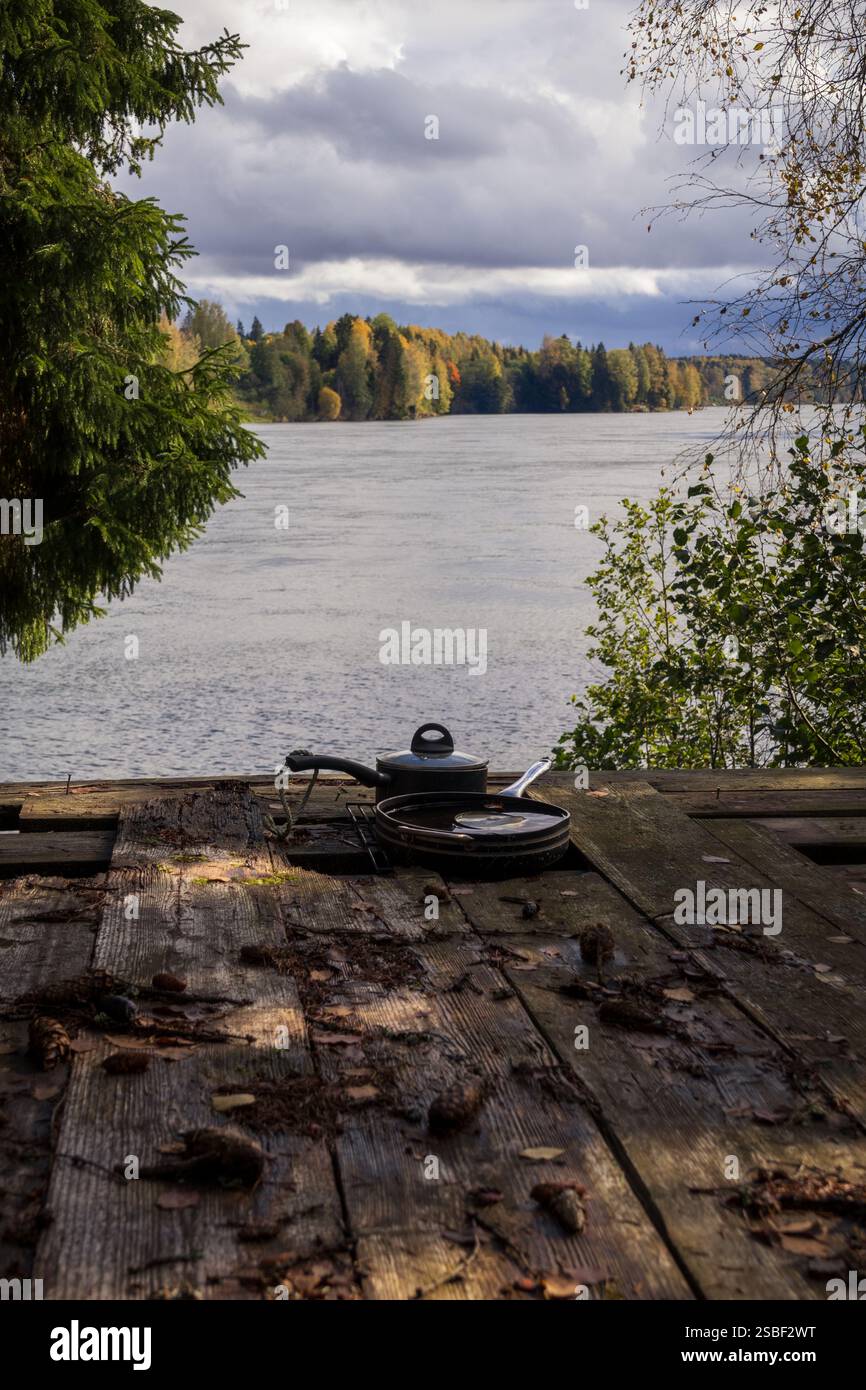 Ein ruhiger Moment am Fluss – Reflektionen im Wald, eine Holzterrasse und vergessene Kochutensilien, die auf eine Geschichte hinweisen, die in der Umarmung der Natur zurückgeblieben ist Stockfoto