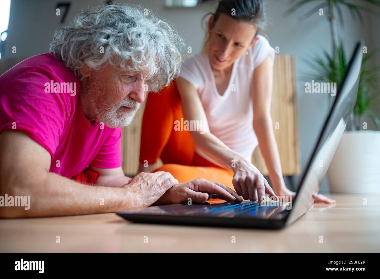 Eine junge Frau führt einen älteren Mann geduldig mit einem Laptop durch und zeigt auf den Bildschirm, während er aufmerksam in einer entspannten Atmosphäre lernt. Stockfoto