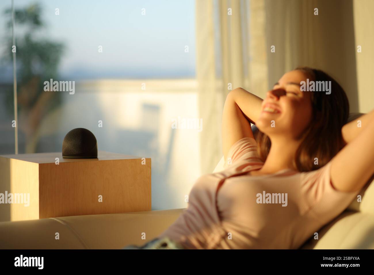 Frau, die sich zu Hause bei Sonnenuntergang mit intelligentem Lautsprecher auf einer Couch entspannt Stockfoto