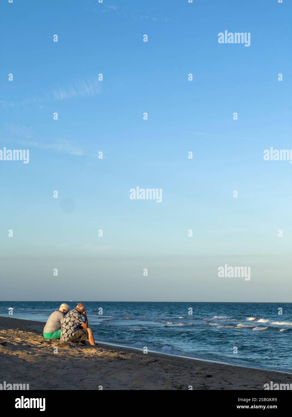 Ein älteres Ehepaar sitzt am Sandstrand und blickt auf die sanften Meereswellen unter dem goldenen Licht am späten Nachmittag in Lauderdale by the Sea, Florida - Smartphone-aufgenommenes Stockfoto