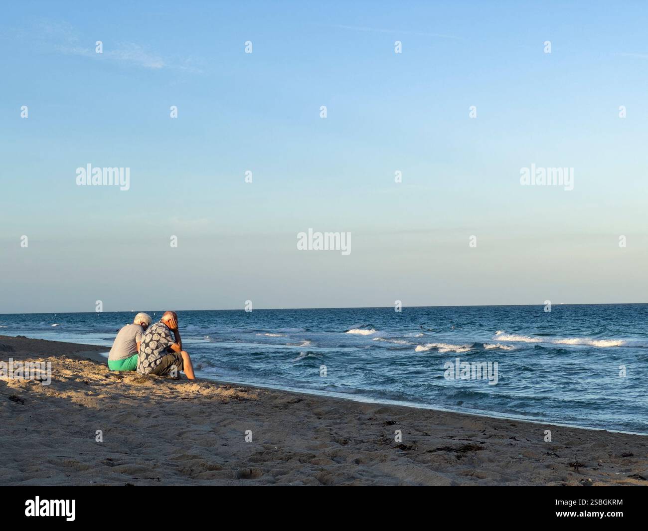 Ein älteres Ehepaar sitzt am Sandstrand und blickt auf die sanften Meereswellen unter dem goldenen Licht am späten Nachmittag in Lauderdale by the Sea, Florida - Smartphone-aufgenommenes Stockfoto