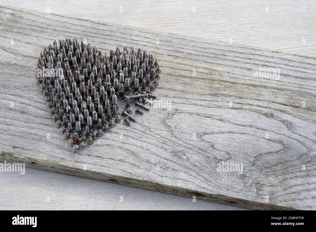 Herzform von kleinen Metallschrauben, die auf ihren Köpfen platziert sind, Schulter an Schulter stehend, eine Seite gebrochen. Hintergrund des verwitterten Holzes. Stockfoto