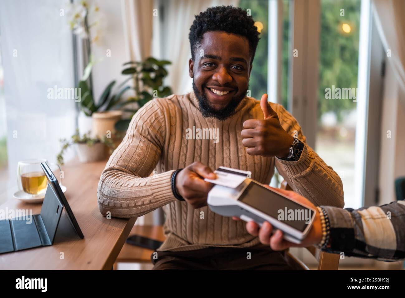 Junger Mann in lässiger Kleidung bezahlt kontaktlos mit Kreditkarte, während er im gemütlichen Stadtcafé sitzt. Stockfoto