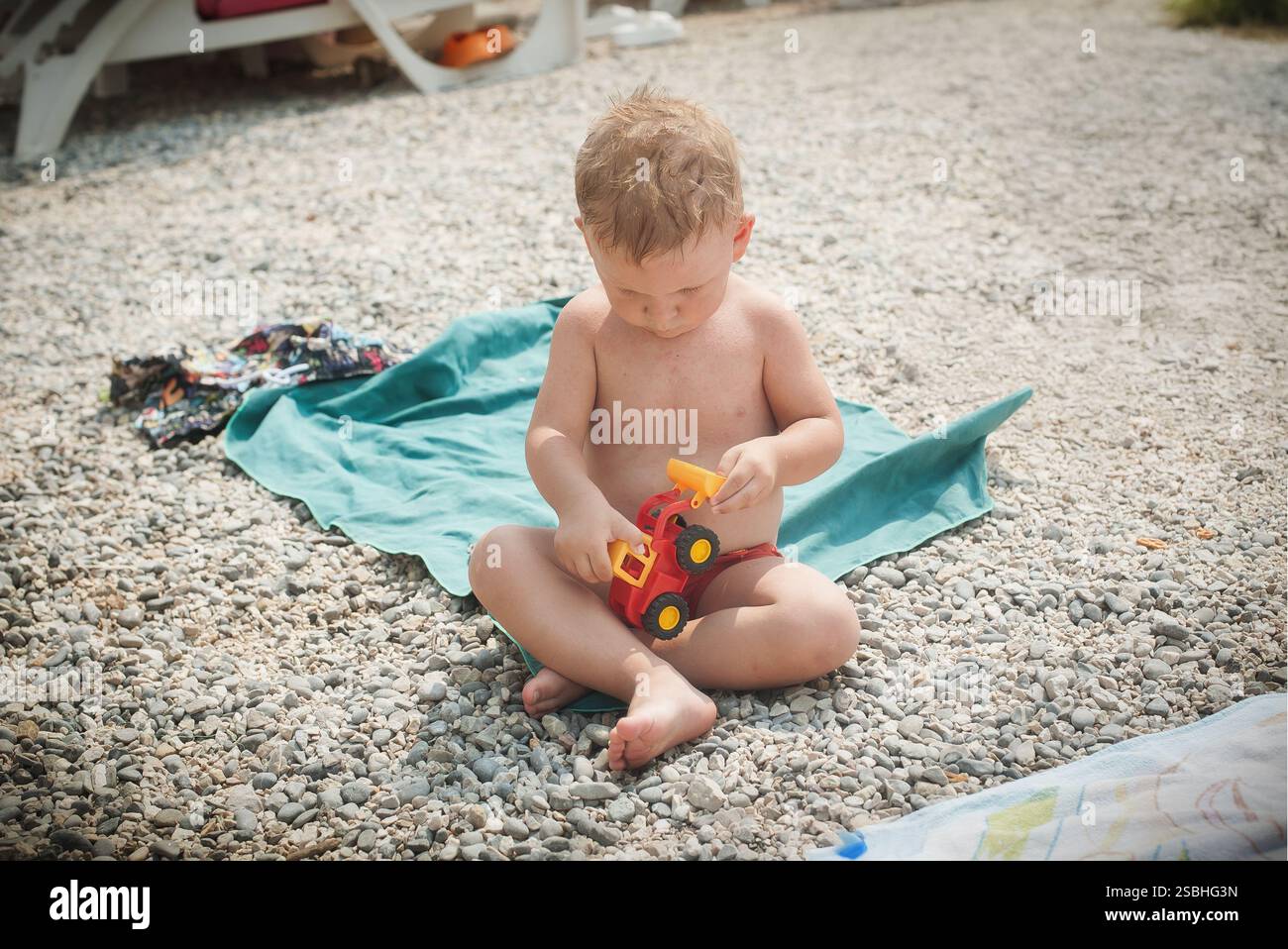 Ein kleines Kind sitzt auf einem grünen Handtuch und spielt mit einem kleinen roten Spielzeugwagen. Die Kiesoberfläche umgibt das Kind und sorgt so für einen verspielten Sommernachmittag Stockfoto