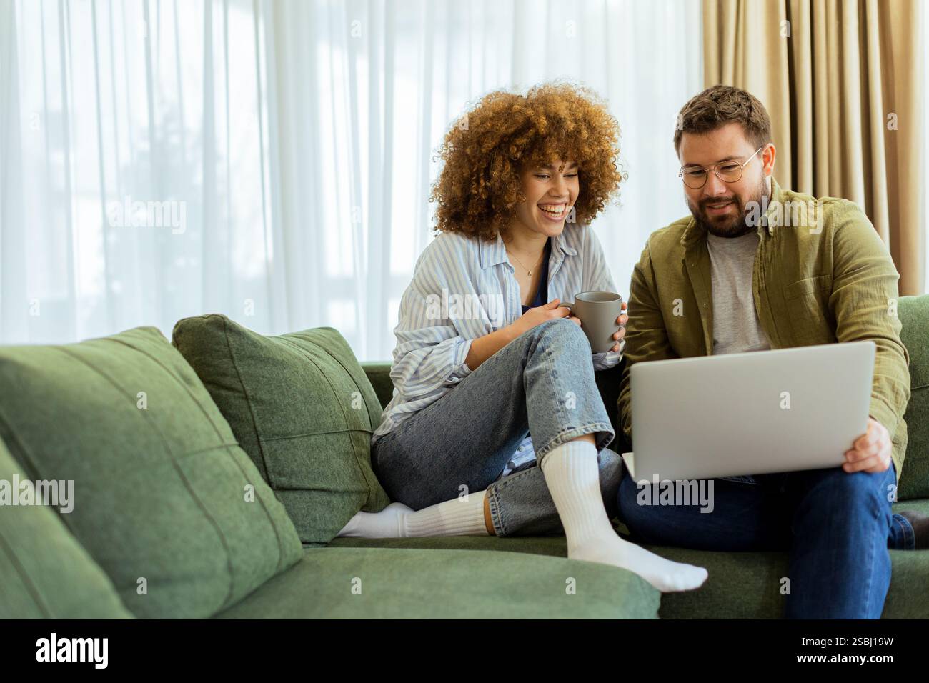 Ein fröhliches Paar sitzt nebeneinander auf einer weichen grünen Couch und führt ein aufregendes Gespräch, während es unter warmem Su auf einem Laptop Ideen durchstöbert Stockfoto