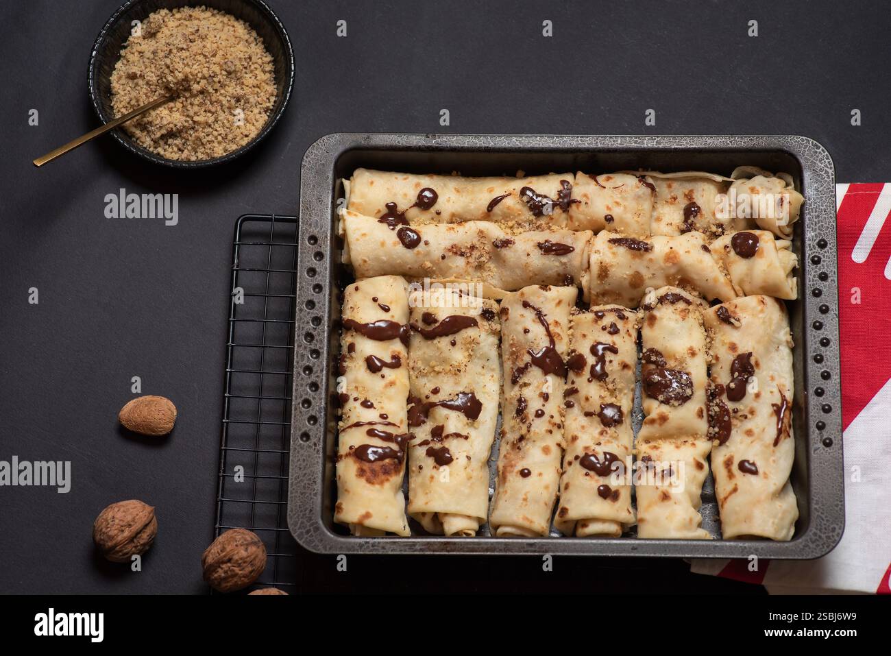 Ein verführerisches Tablett aus frisch gebackenen, mit Schokolade bespritzten Crepes, garniert mit fein gehackten Nüssen, platziert vor einem eleganten schwarzen Hintergrund, das Kuli zeigt Stockfoto