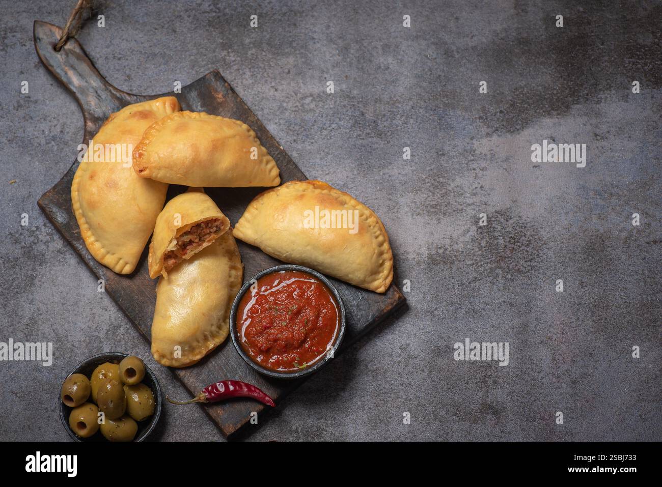Dieses Bild zeigt köstliche goldene, frittierte Empanadas, die auf einem hölzernen Schneidebrett angeordnet sind, begleitet von einer Schüssel mit reichhaltiger Tomatensalsa und dekorativem Dekor Stockfoto
