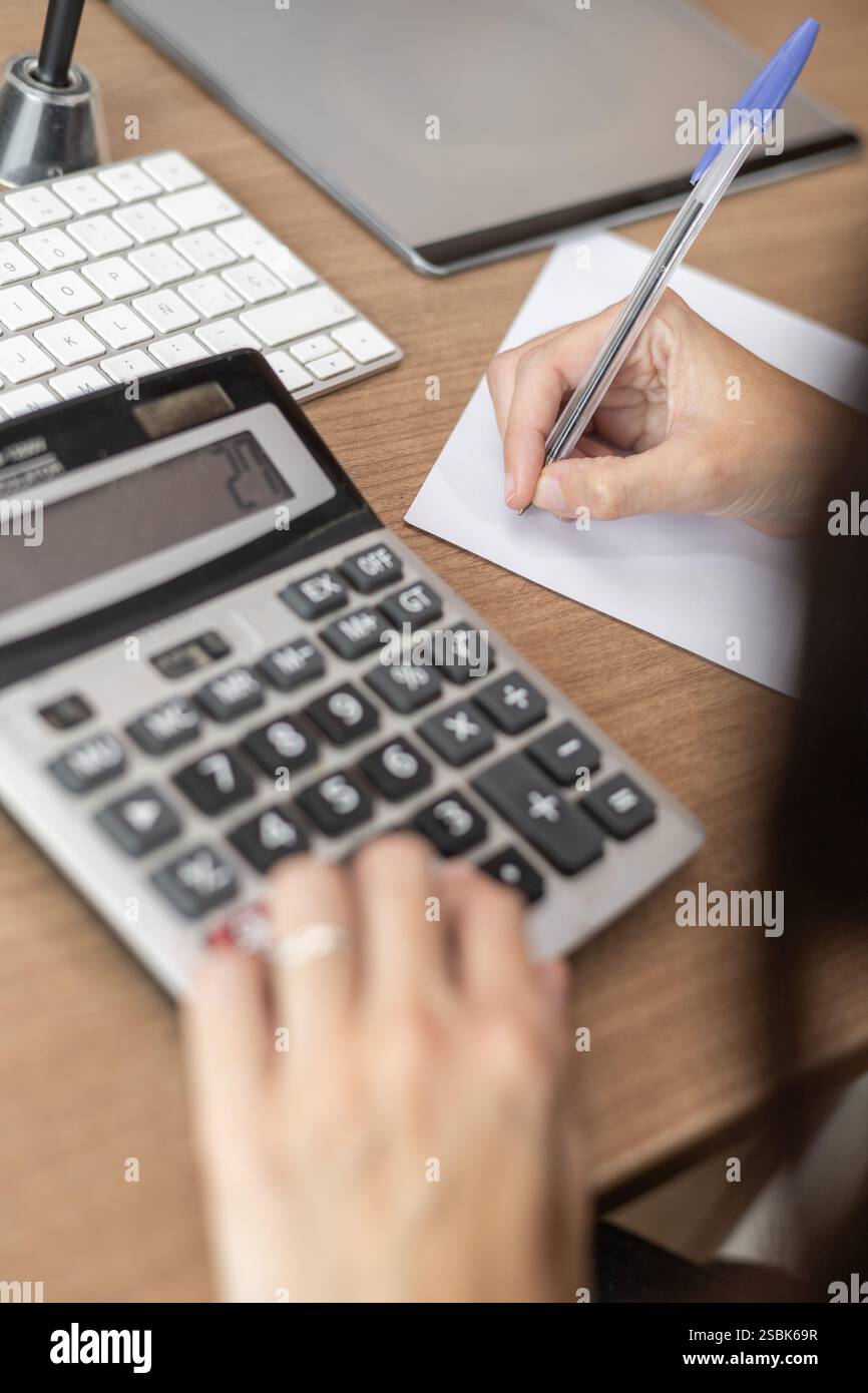 Eine fokussierte Frau, die Berechnungen an einem Rechner durchführt und mit einem Stift auf ein Blatt Papier schreibt. Perfekt für Finanzen, Bildung, Produktivität und Stockfoto