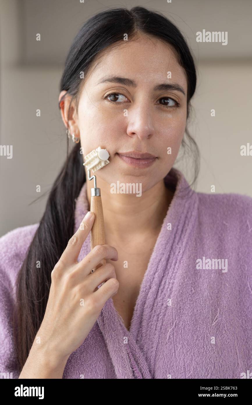 Eine schöne Frau in einem rosafarbenen Bademantel, die Creme auf ihr Gesicht aufträgt und ihre Hautpflegeroutine verbessert. Perfekt für Beauty-, Self-Care- und Wellness-Inhalte. Stockfoto