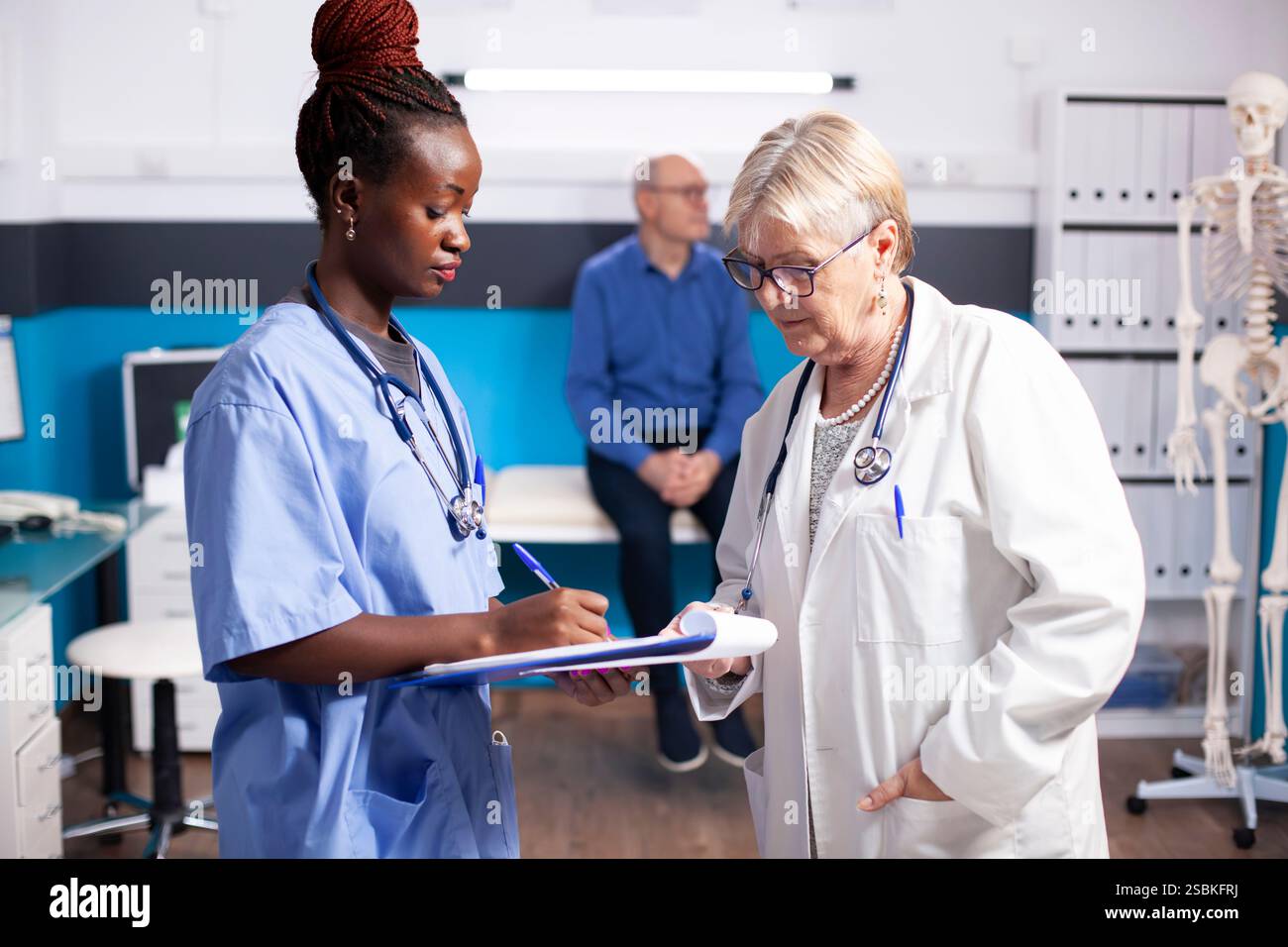 afroamerikanische Krankenschwester hält Klemmbrett und prüft medizinische Notizen mit einem leitenden Arzt, der im Klinikzimmer steht. Diverse Krankenhauspersonal diskutieren als alter männlicher Patient im Hintergrund. Stockfoto