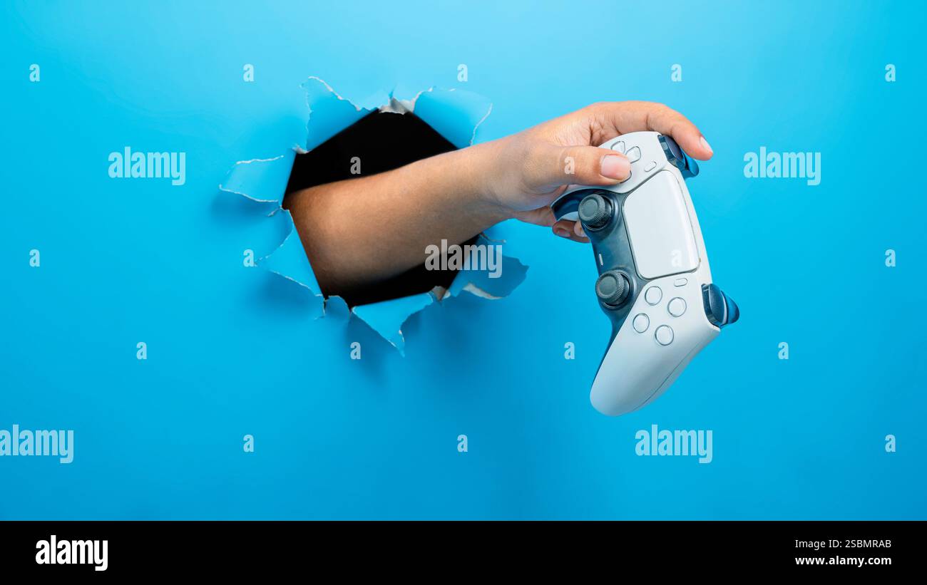Menschliche Hand hält Gamepad, Joystick oder Videospiel-Controller durch gerissenes Papier. modellerstellung oder Kopierbereich. Konzept des Spielens von Videospielen, Enterta Stockfoto