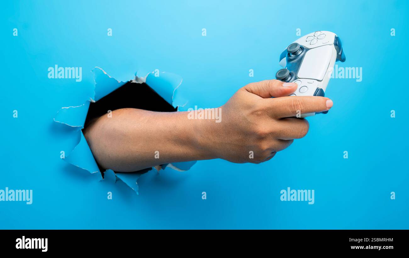 Menschliche Hand hält Gamepad, Joystick oder Videospiel-Controller durch gerissenes Papier. modellerstellung oder Kopierbereich. Konzept des Spielens von Videospielen, Enterta Stockfoto