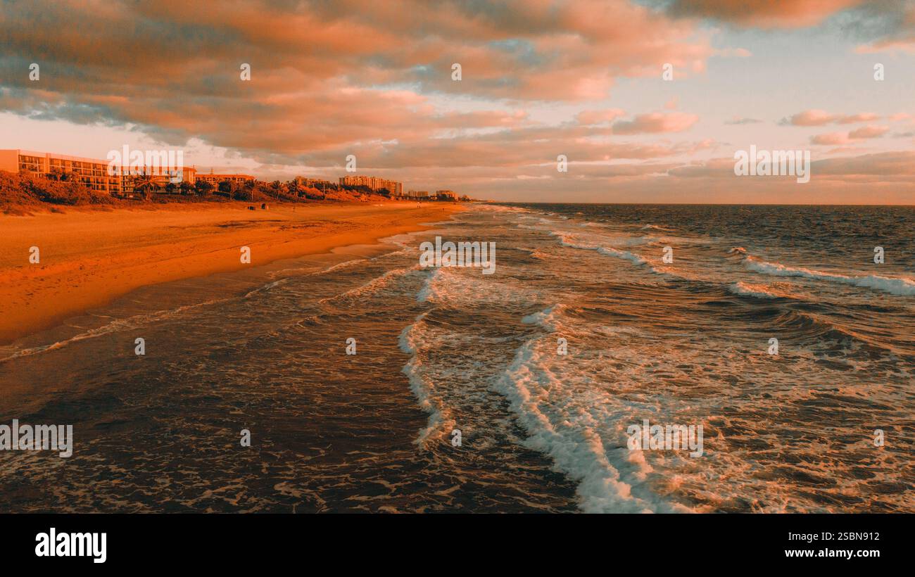 Sanfte Wellen Rollen auf einen ruhigen Strand, während die Sonne untergeht, und schaffen einen atemberaubenden goldenen Farbton über dem Wasser und Sand. Wolken verleihen dem farbenfrohen Himmel Tiefe. Stockfoto