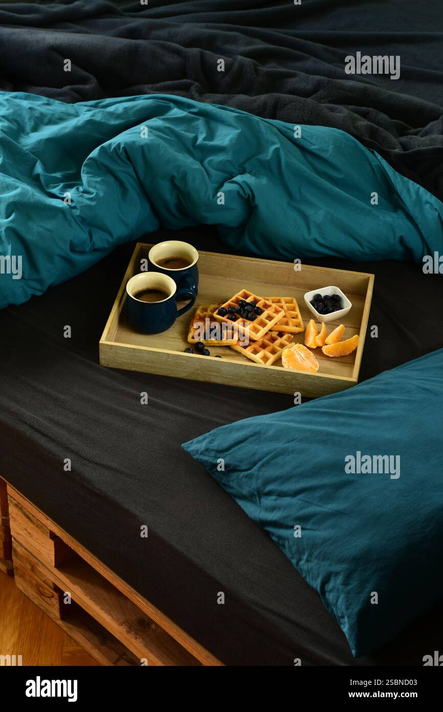 Waffeln, Kaffee, Blaubeeren und Orangenfrühstück auf einem Tablett im Holzpalettenbett auf schwarzen und dunkelblauem Laken Stockfoto
