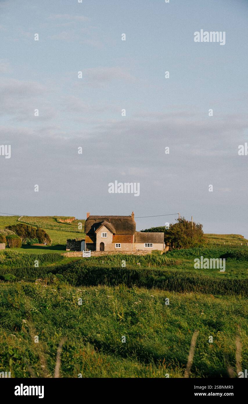 Ein kleines Haus sitzt auf einem Grasfeld. Der Himmel ist bewölkt und die Sonne scheint Stockfoto