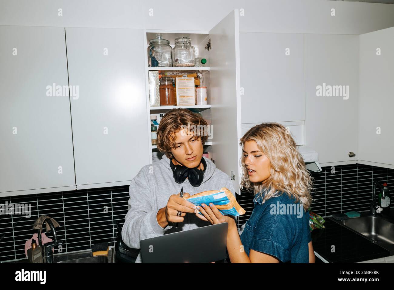 Junge männliche und weibliche Freunde lesen Details auf der Essenspackung, während sie in der Küche des Schlafsaalzimmers stehen Stockfoto