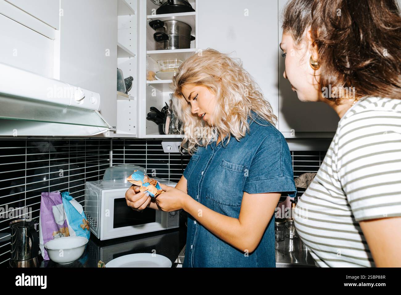 Blonde Frau untersucht das Essenspaket, während sie mit einer Freundin in der Küche des Studentenwohnheims steht Stockfoto