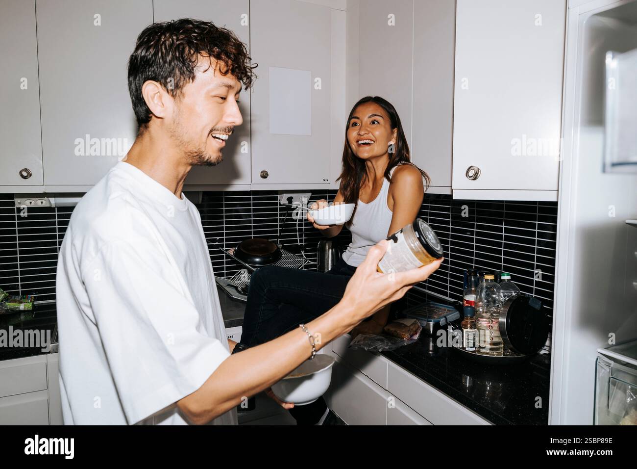 Lächelnder Mann, der das Lebensmitteletikett überprüft, während er mit einer weiblichen Mitbewohnerin im Studentenwohnheim auf der Küchenzeile redet Stockfoto