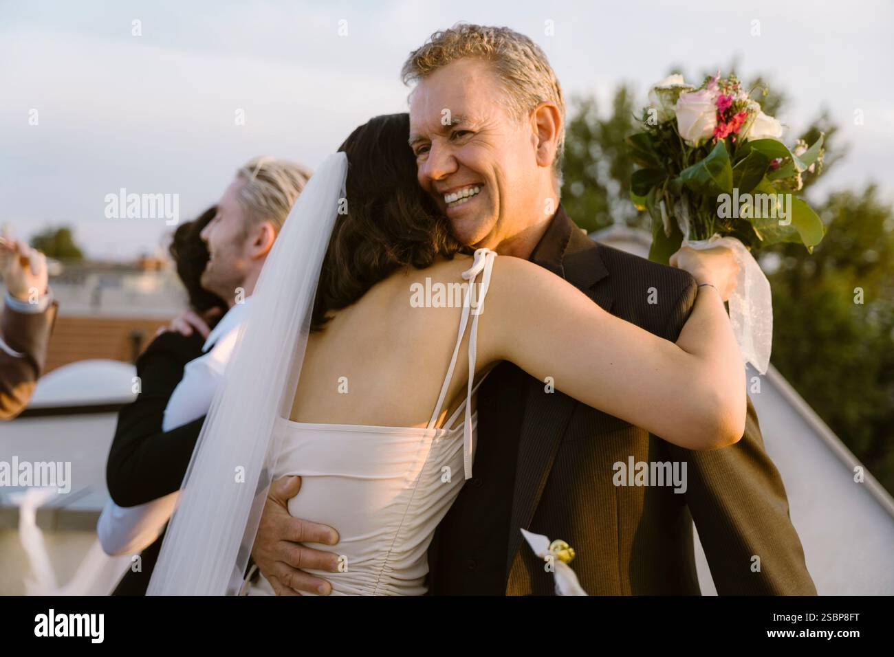 Glücklicher Senior-Mann, der die frisch vermählte Tochter umschließt und beim Hochzeitsempfang einen Blumenstrauß hält Stockfoto