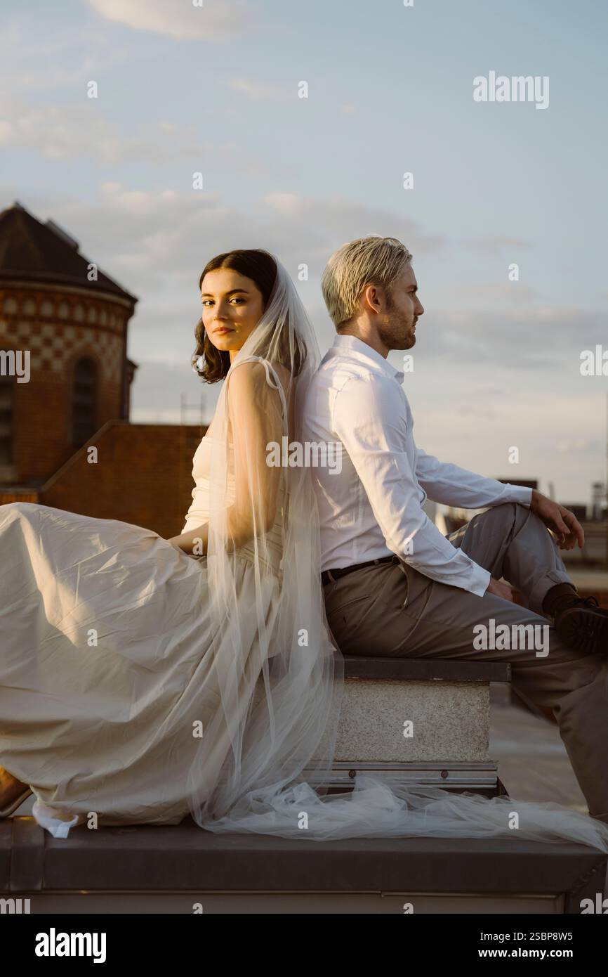 Seitliches Porträt eines jungen, frisch verheirateten, multirassischen Paares, das auf dem Dach gegen den Himmel sitzt Stockfoto