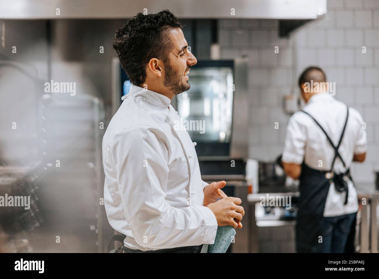 Seitenansicht eines glücklichen jungen männlichen Küchenchefs in weißer Uniform, der in der Restaurantküche steht Stockfoto