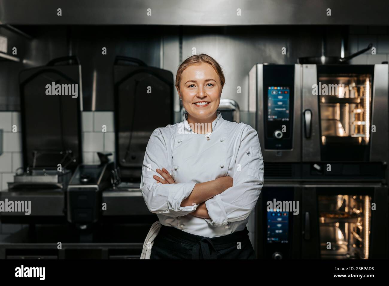 Porträt einer lächelnden Küchenchefin in weißer Uniform und stehend mit überkreuzten Armen in der Küche im Restaurant Stockfoto