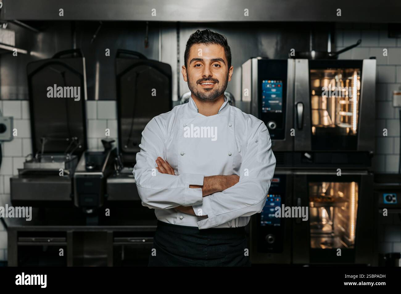 Porträt eines selbstbewussten jungen Küchenchefs in weißer Uniform und stehend mit überkreuzten Armen in der Küche des Restaurants Stockfoto