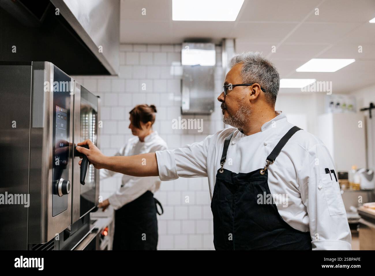 Reifer männlicher Koch, der Schürze mit Backofen in der kommerziellen Küche trägt Stockfoto