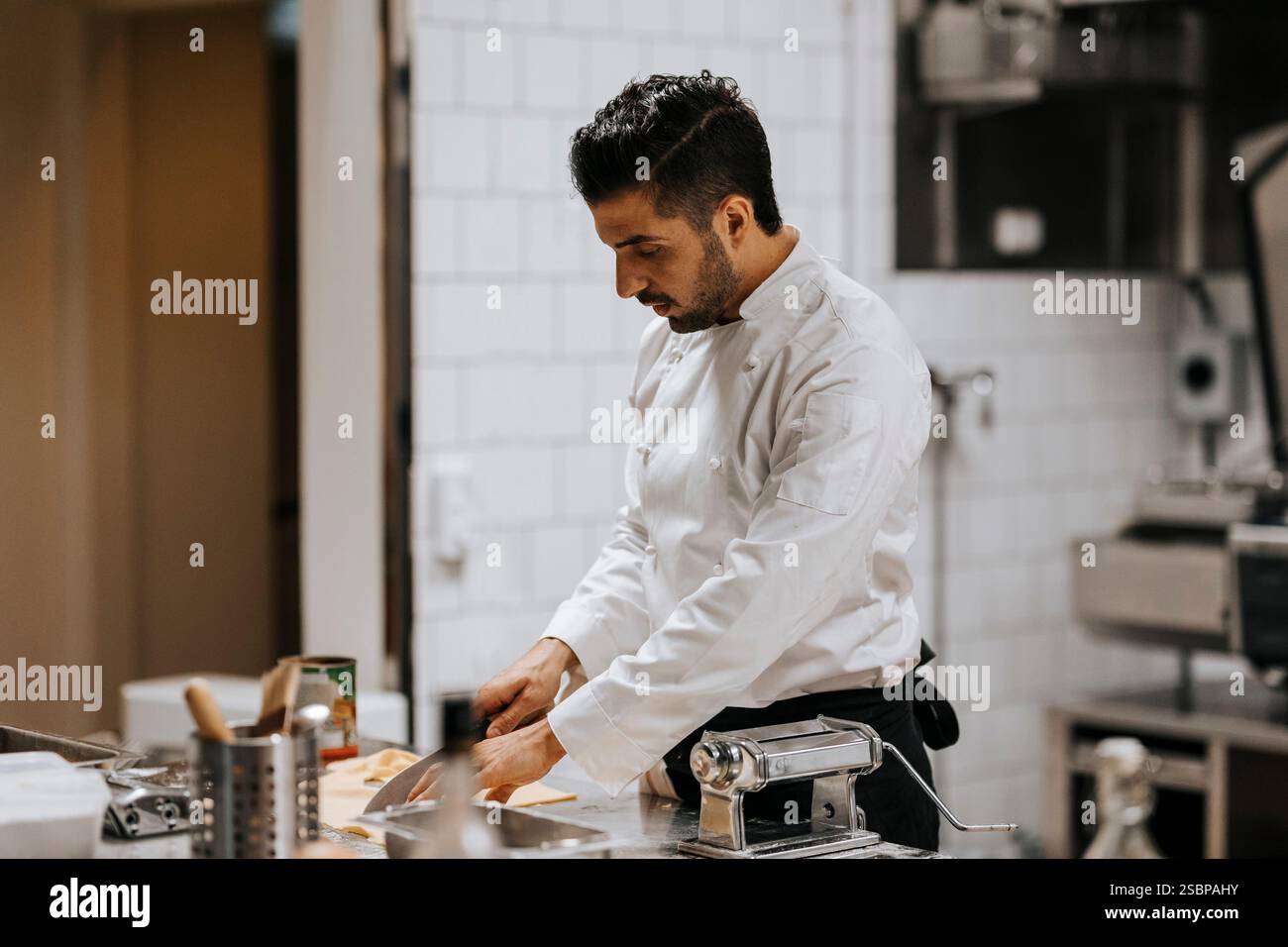 Konzentrierter junger männlicher Koch in weißen Uniformen, der auf der Küchentheke im Restaurant hackt Stockfoto