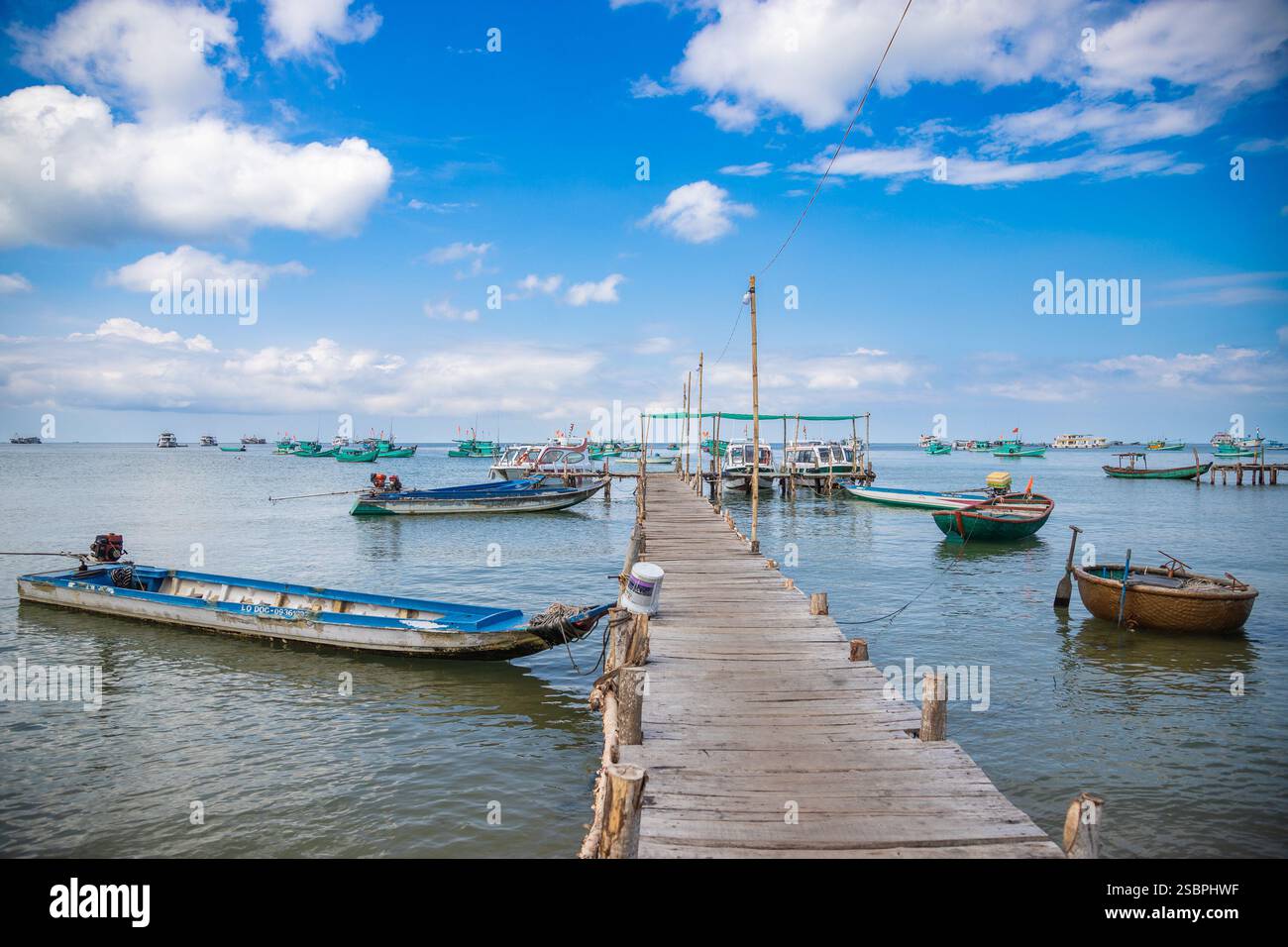 Eine friedliche Küstenlandschaft mit einem hölzernen Pier, der sich bis zum Horizont erstreckt, umgeben von traditionellen Booten, die auf ruhigem, flachem Wasser ankern. Stockfoto