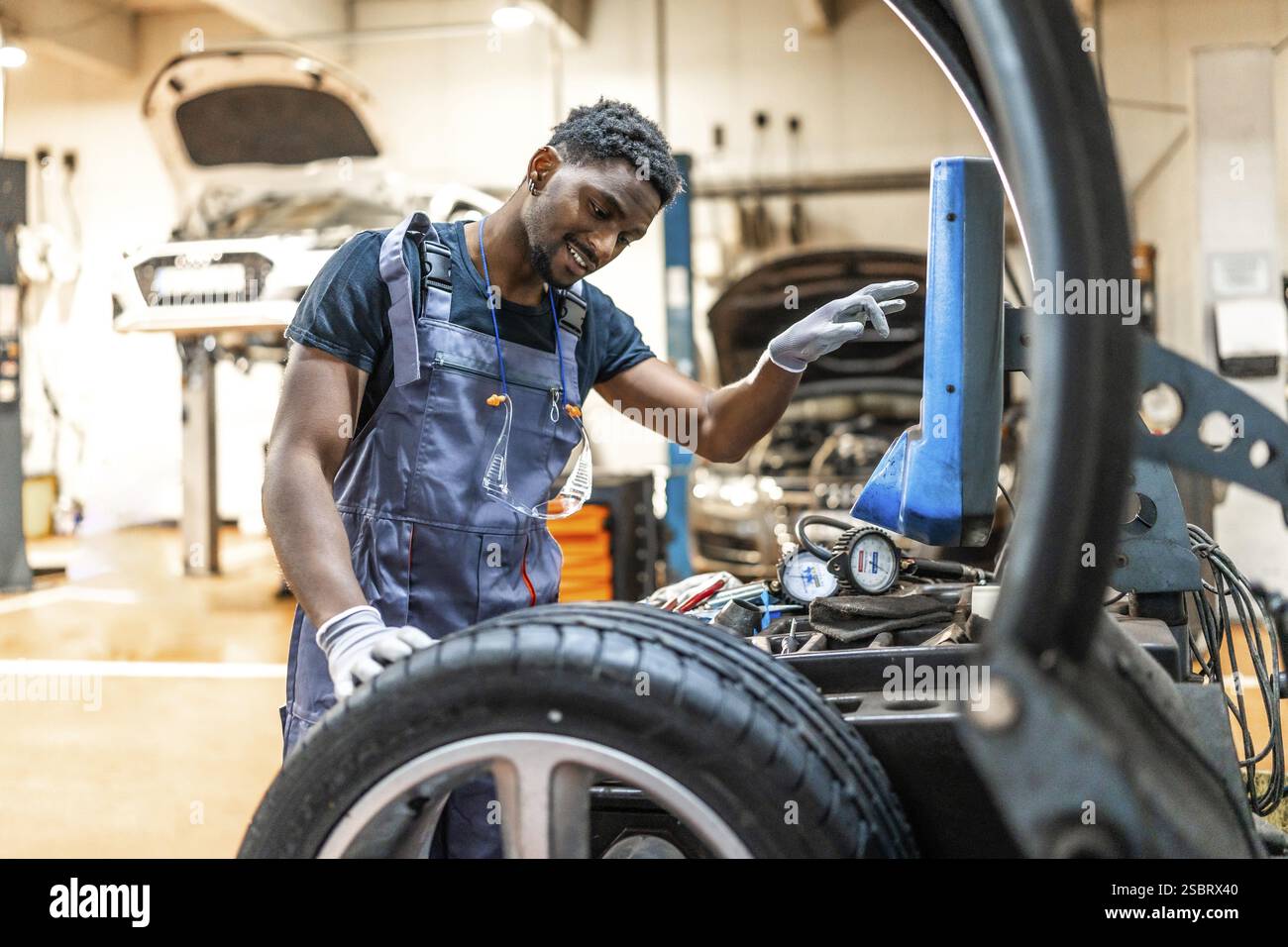 Junger, lächelnder afroamerikanischer Mechaniker, der Autoreifen mit einem digitalen Reifenwechsler in einer Autowerkstatt wechselt Stockfoto