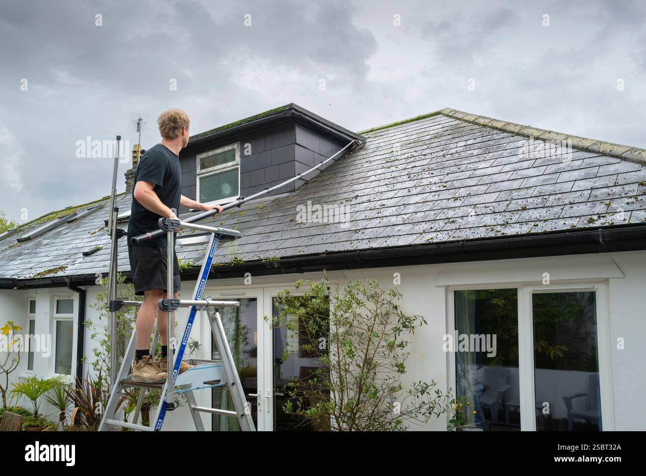 Ein Mann, der auf einer Leiter steht und Moos von einem Dach eines Hauses in Newquay in Cornwall in Großbritannien reinigt. Stockfoto