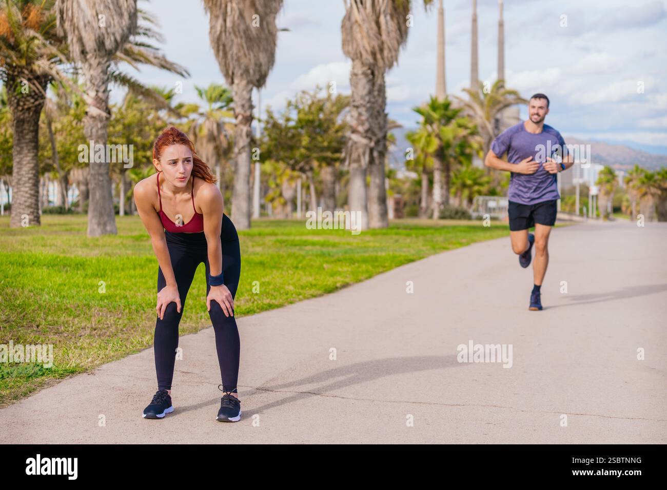 Müde Frau ruht sich nach dem Joggen aus, während der Mann weiter in einem Park läuft Stockfoto