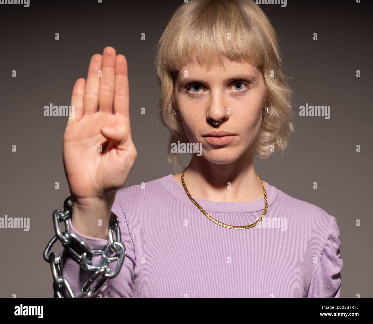 Eine junge Frau mit kurzen blonden Haaren, die einen lila Pullover trägt, macht ein leises Handzeichen für Hilfe, während ihr Handgelenk in Metallketten gewickelt ist, Symbol Stockfoto