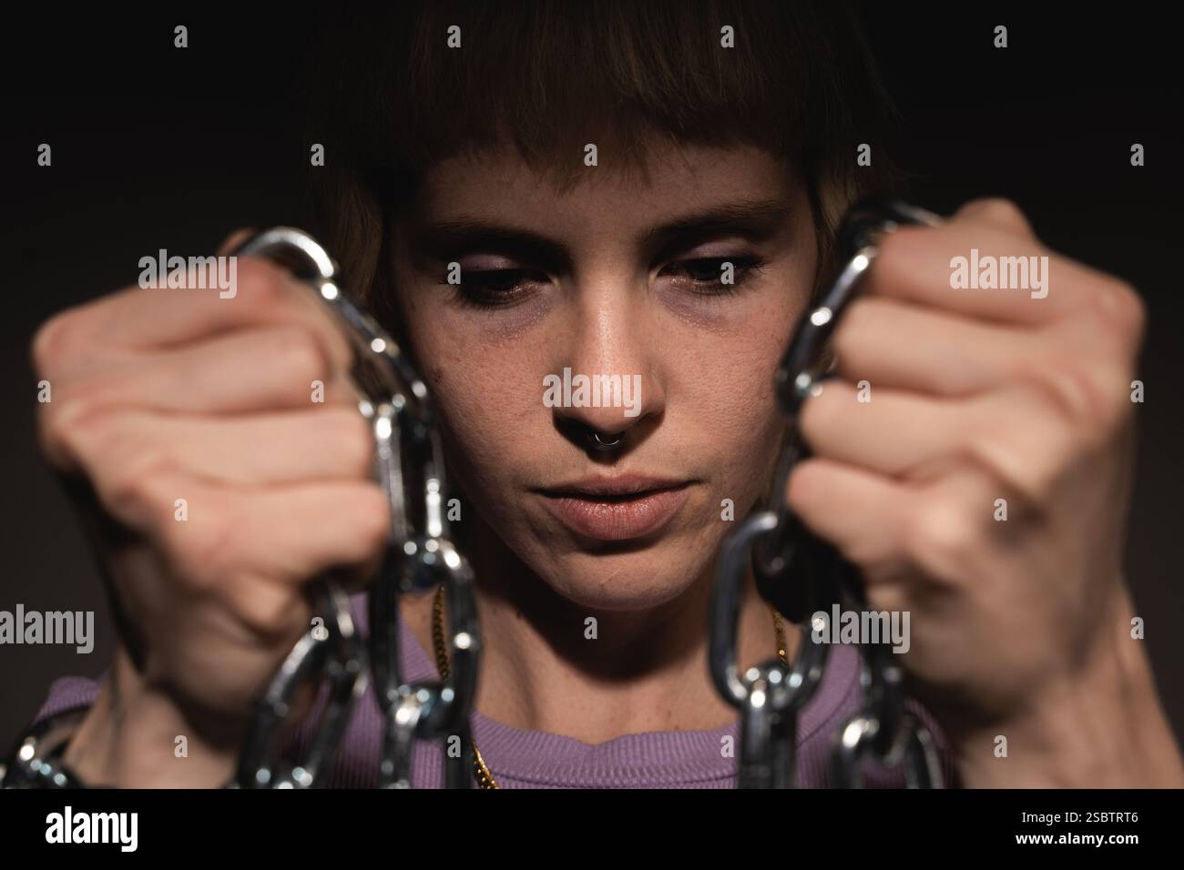 Eine junge Frau mit kurzen blonden Haaren, die einen lila Pullover trägt, greift Metallketten und symbolisiert Unterdrückung, geschlechtsspezifische Gewalt und den Kampf für die Frauen Stockfoto