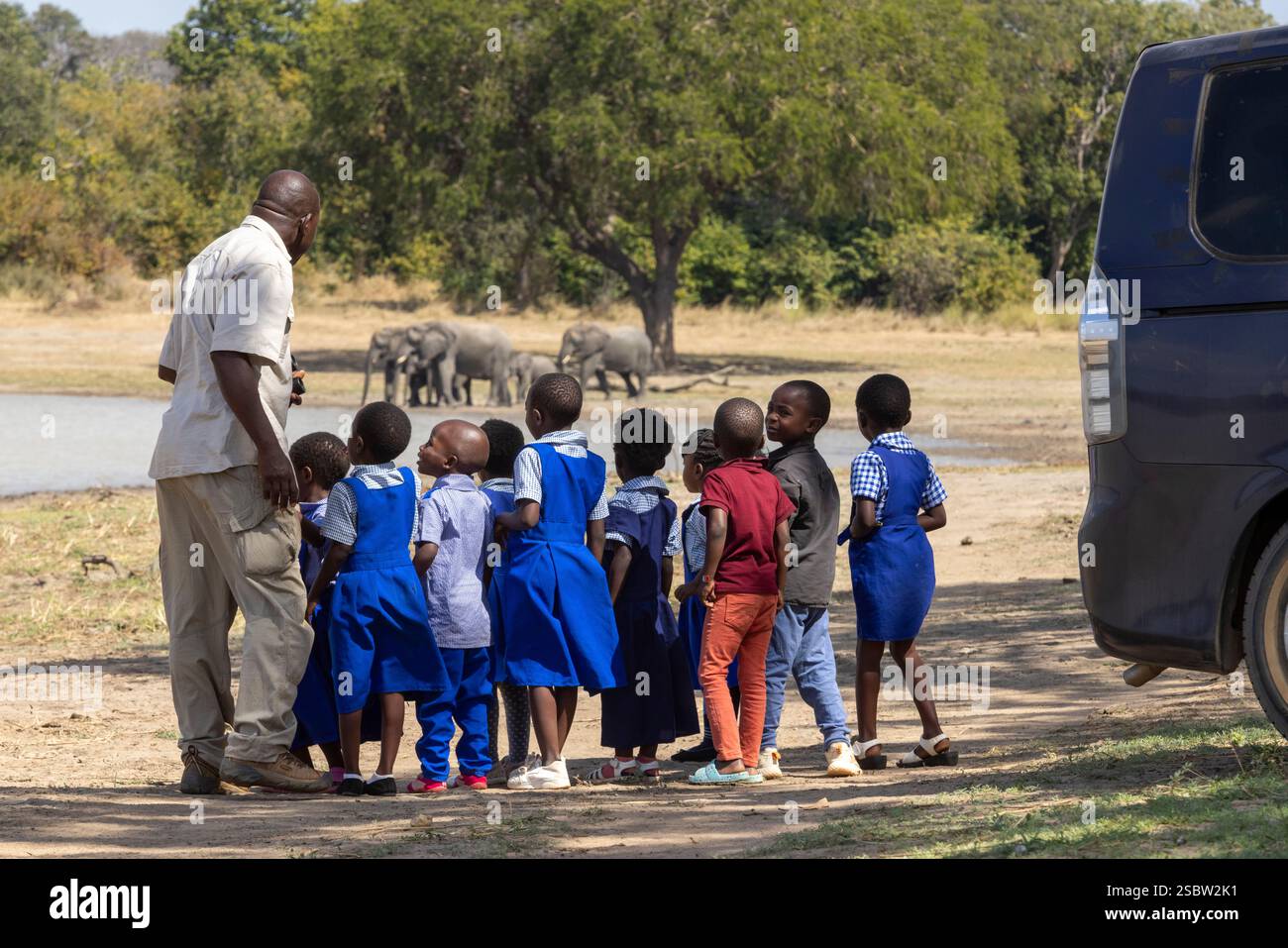 Eine Gruppe von Schulkindern auf einem Ausflug, die eine entfernte Elefantenherde beim Trinken im Vwaza Marsh Wildlife Reserve beobachten Stockfoto