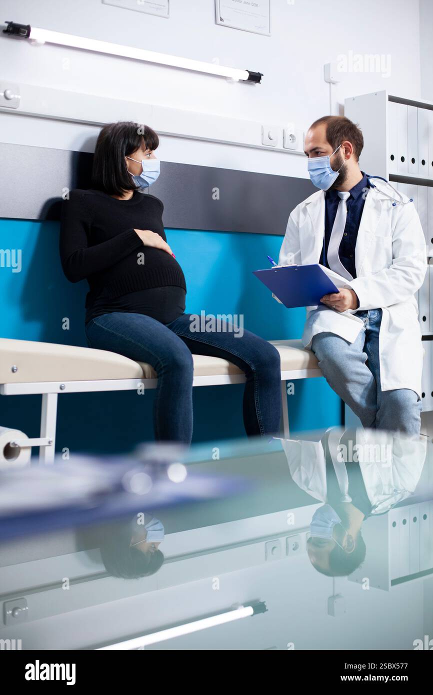 Die Patientin in der Schwangerschaftsbetreuung sitzt neben dem Arzt im Labormantel und bietet Untersuchungsergebnisse und pränatale Pflegeoptionen. Arzt hält Clipboard und überprüft ärztliche Anweisungen bei schwangeren Frauen. Stockfoto