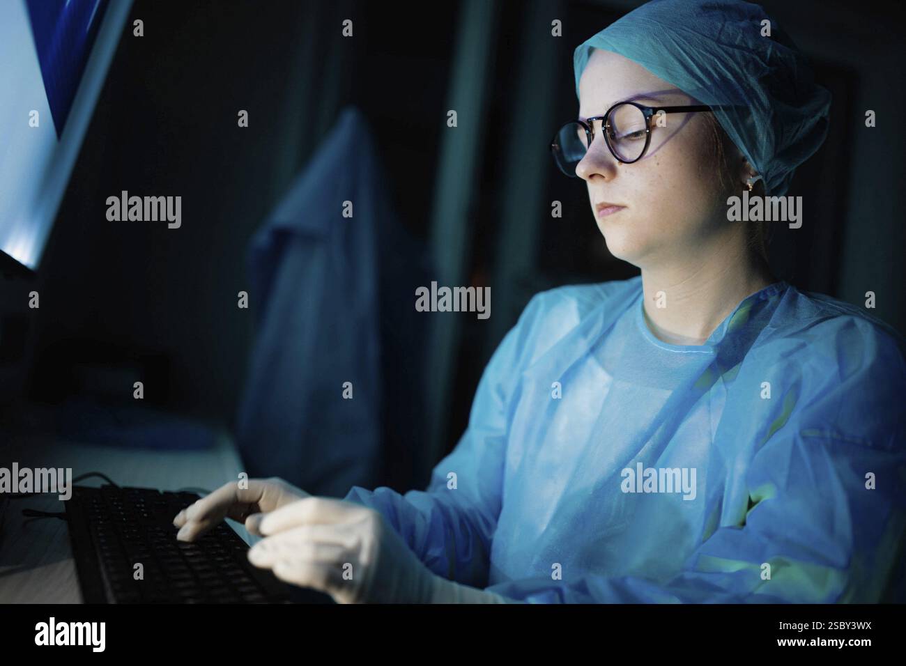 Ein medizinischer Mitarbeiter in blauem Kleid, der an einem Computer bei schwacher Beleuchtung arbeitet Stockfoto