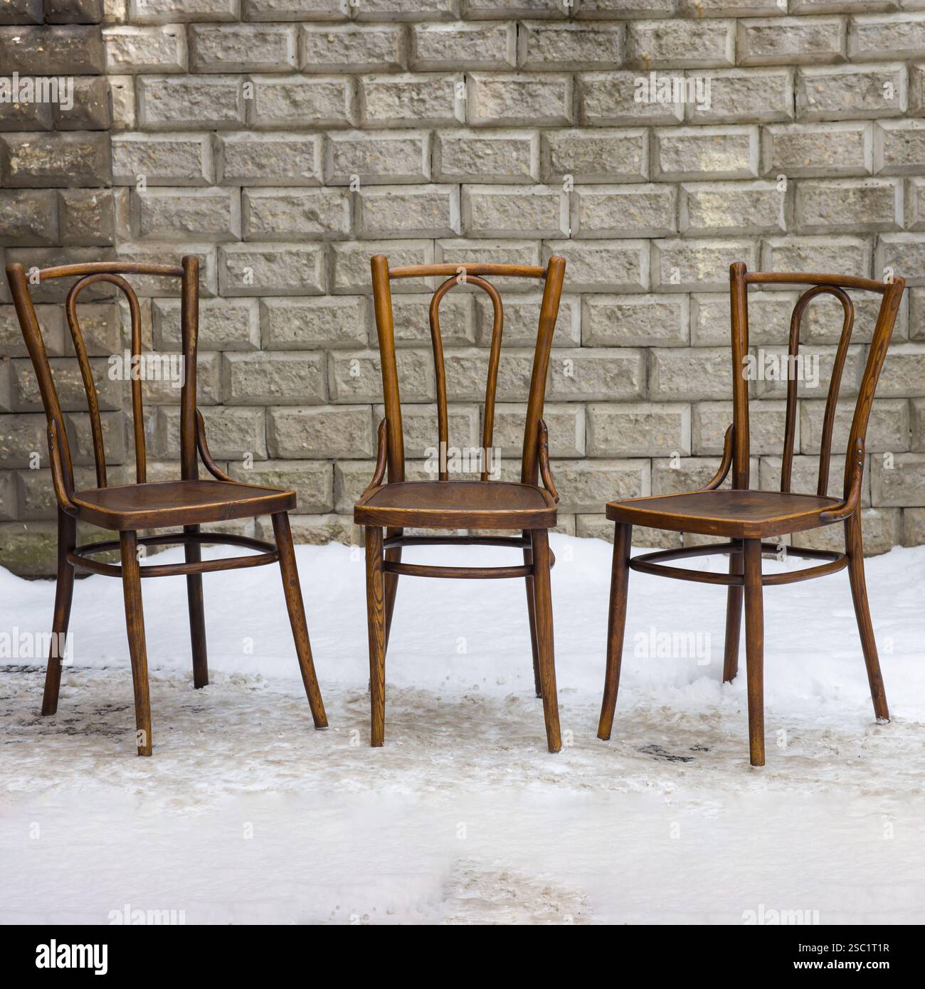 Drei Vintage-Bugholzstühle, elegant gefertigt und in Braun gehalten, stehen nebeneinander an einer strukturierten grauen Ziegelwand auf einer schneebedeckten Oberfläche. Stockfoto