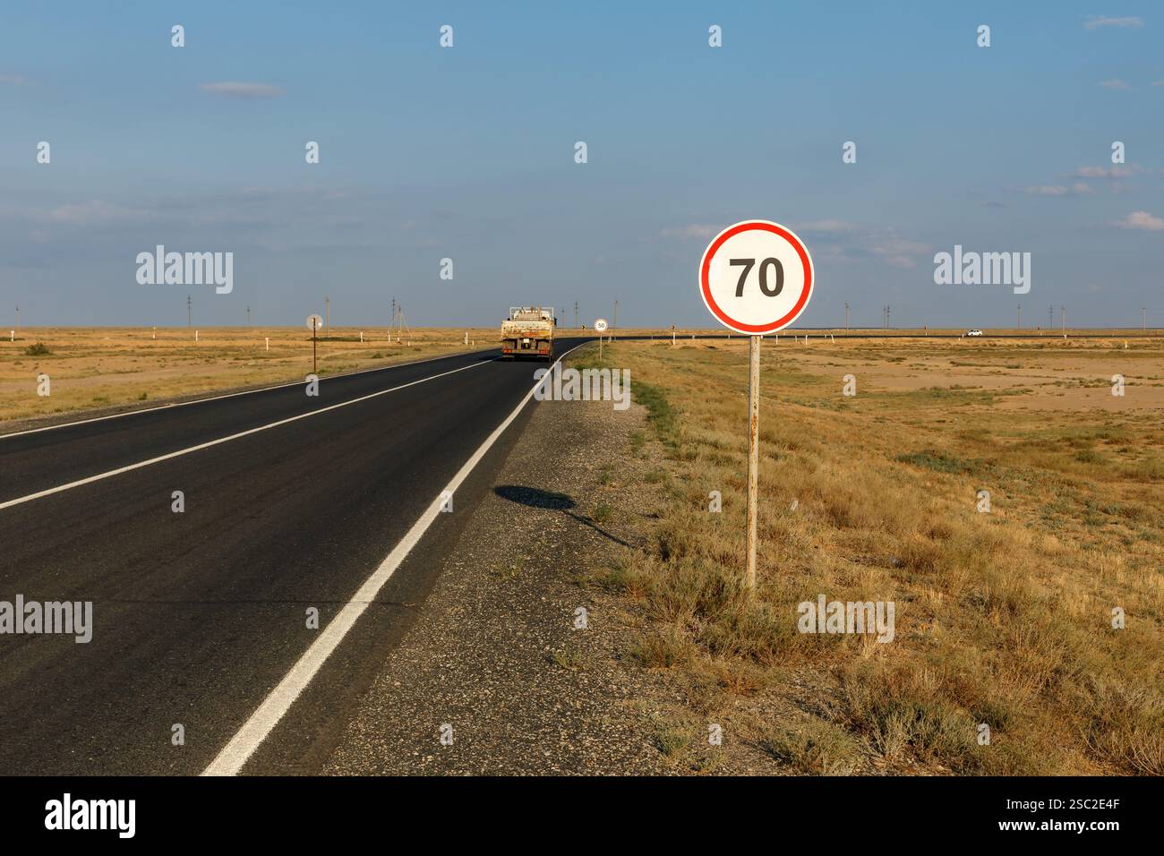 Eine Landstraße führt durch die Region Atyrau in Kasachstan, auf der ein Geschwindigkeitsbegrenzungsschild von 70 km und ein sich näherndes Fahrzeug abgebildet sind. Stockfoto