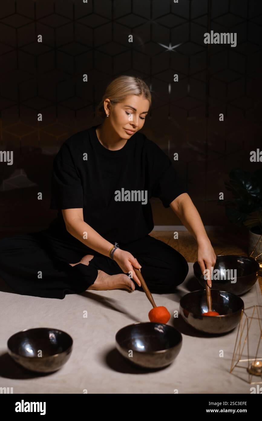 Eine Frau sitzt ruhig auf dem Boden und übt Klangheilung mit mehreren Klangschalen, die um sie herum angeordnet sind. Die Einstellung umfasst sanfte Beleuchtung und pla Stockfoto