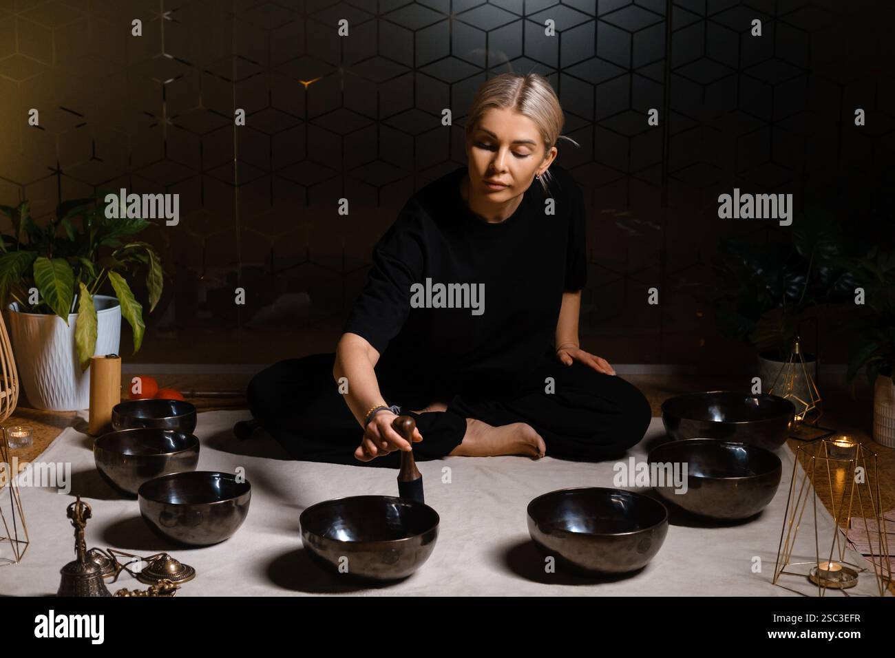 Eine Frau sitzt ruhig auf dem Boden und übt Klangheilung mit mehreren Klangschalen, die um sie herum angeordnet sind. Die Einstellung umfasst sanfte Beleuchtung und pla Stockfoto