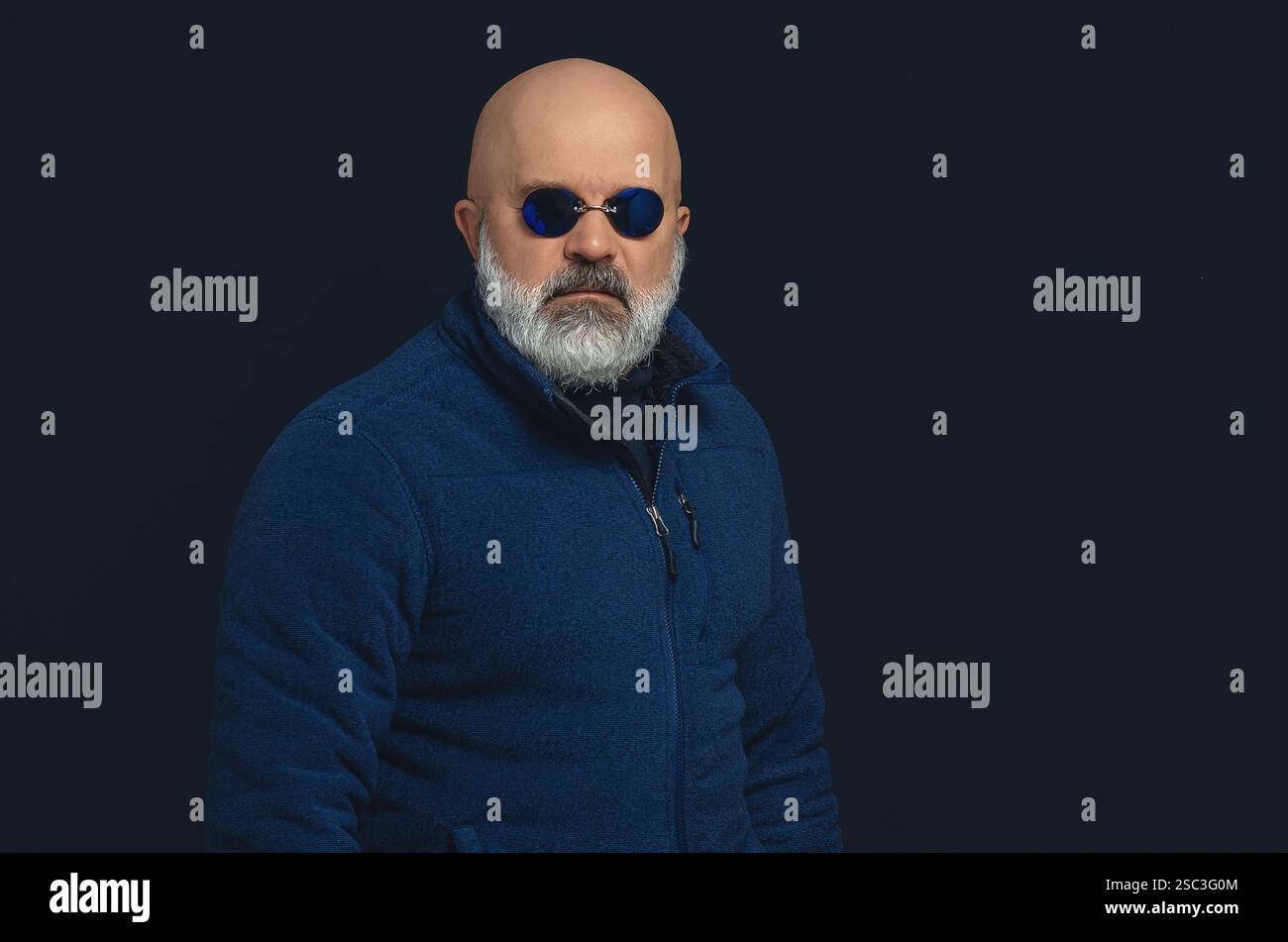 Glatzbärtiger brutaler Mann in dunkelblauem Pullover und Brille. Blick auf die Kamera. Schwarzer Studiohintergrund Stockfoto